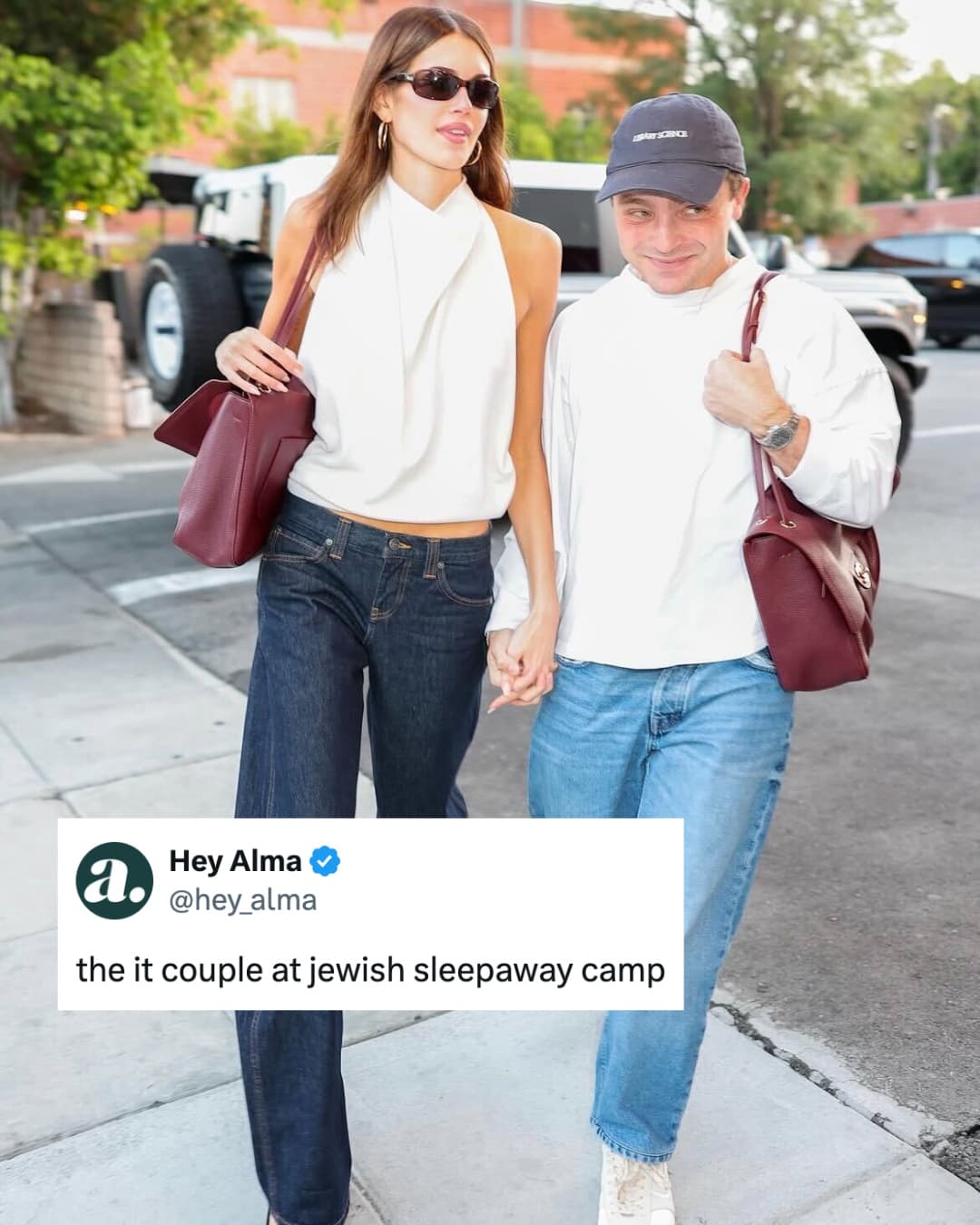 A paparazzi photograph of a young couple walking hand in hand on a pavement, both in white tops and jeans and each carrying a matching burgundy leather handbag, captioned by a Hey Alma post reading 'the it couple at jewish sleepaway camp'.